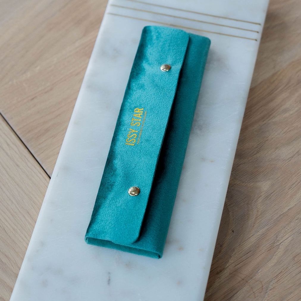 A teal velvet jewellery pouch with gold snap buttons and gold 'ISSY STAR' branding, displayed on a white marble tray over a light wood background.