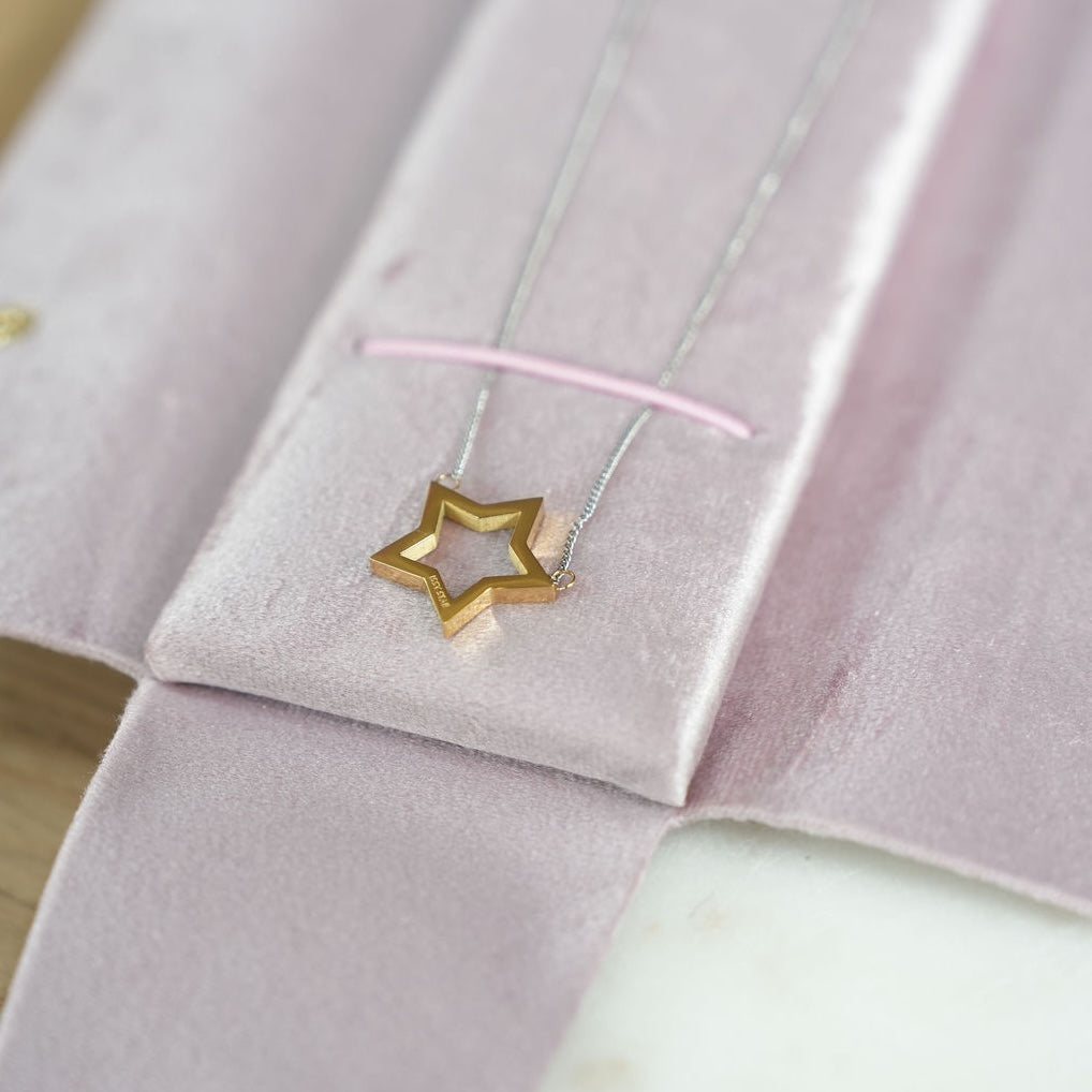 A gold star-shaped pendant necklace with a silver-toned chain, in a light pink velvet jewellery pouch. 